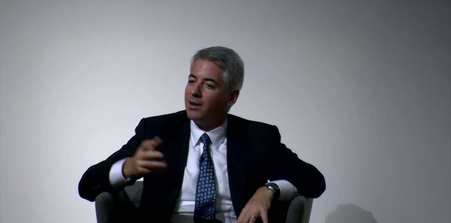 A man in a dark suit and patterned tie sits in a chair against a plain background, gesturing with his left hand while looking slightly to the side.