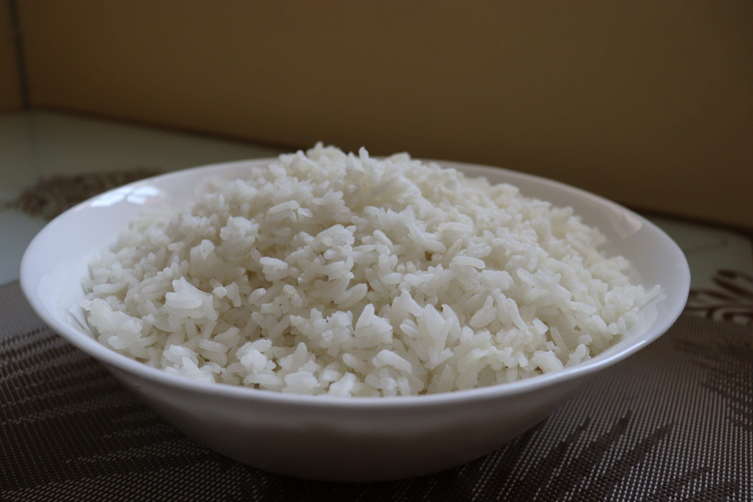 A white bowl filled with a mound of cooked white rice sits on a table with a patterned placemat and a light-colored background.