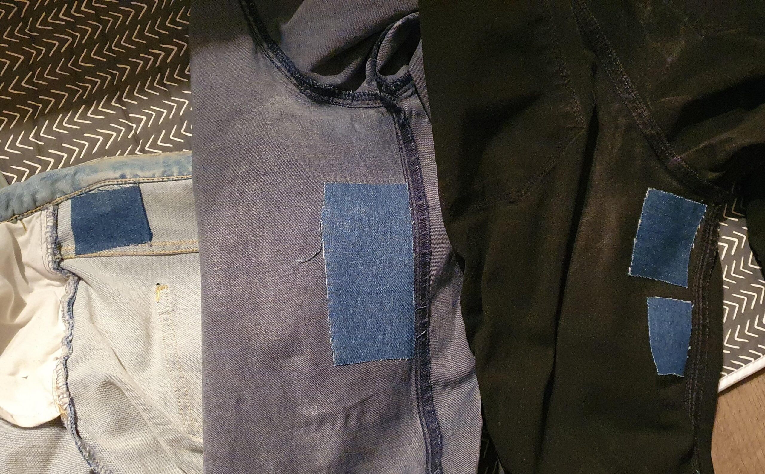 Three pairs of jeans are turned inside out, each showing denim patches sewn onto worn areas to repair holes. The patches are blue and contrast with the original fabric colors. The jeans are laid out on a patterned surface.