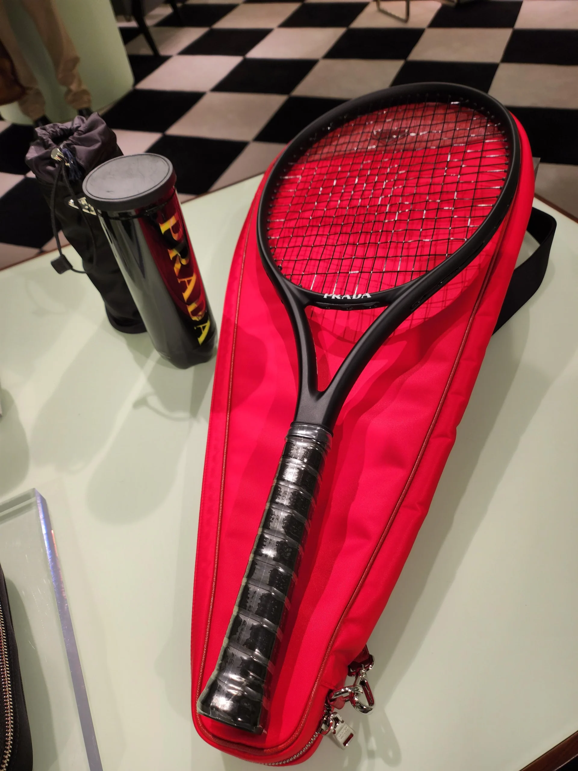 A black tennis racket rests on a bright red case on a table, with a black Prada-branded tennis ball canister nearby. In the background, a checkered black-and-white floor is visible.