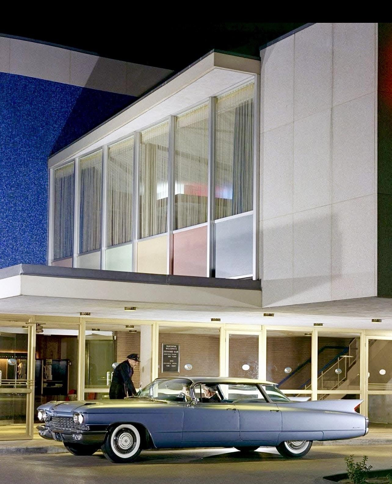 A vintage blue car is parked outside a modern mid-century building at night. A man in a suit stands near the entrance, while another person sits in the car. The building has large glass windows and colorful panels inside.