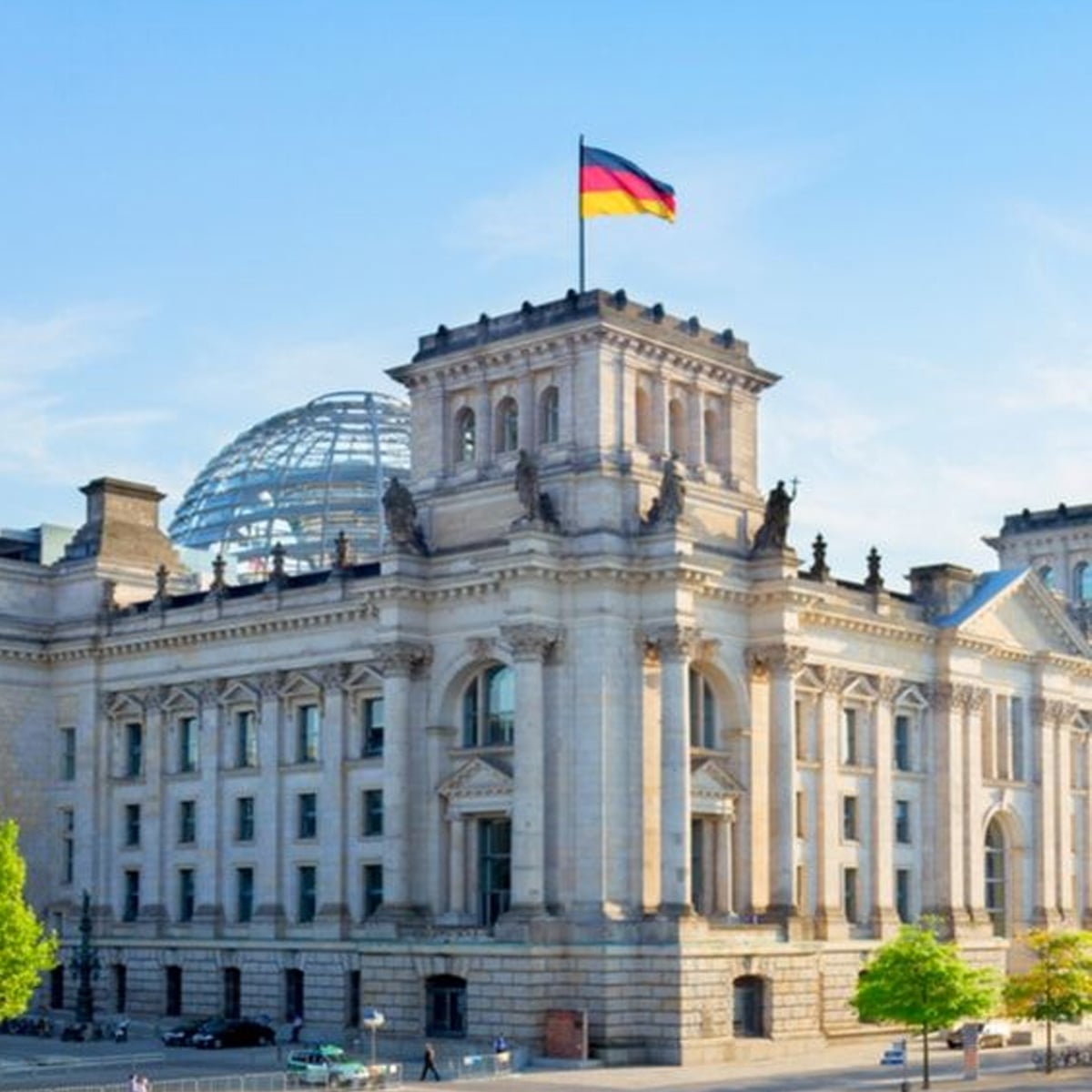 The German Reichstag building in Berlin, featuring a glass dome and the German flag flying on top, under a clear blue sky.