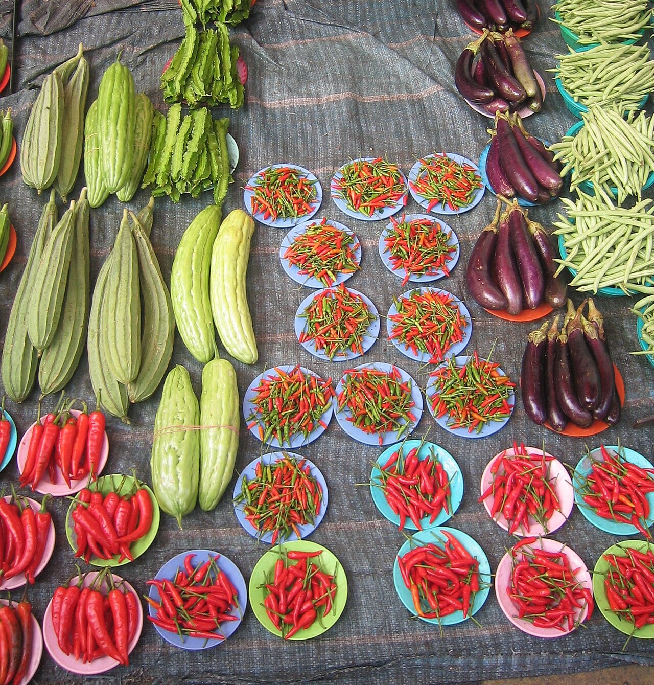 Colorful plates filled with red chili peppers are arranged on a fabric surface, surrounded by green gourds, bitter melons, eggplants, and green beans at a vegetable market.