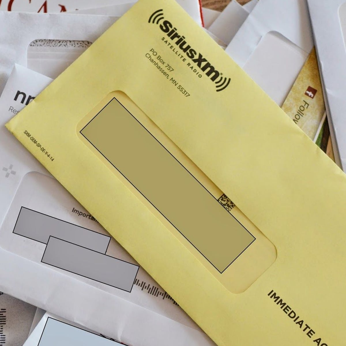 A yellow SiriusXM envelope is stacked on top of several other white envelopes, some with address windows visible. The envelopes appear to be assorted pieces of mail.