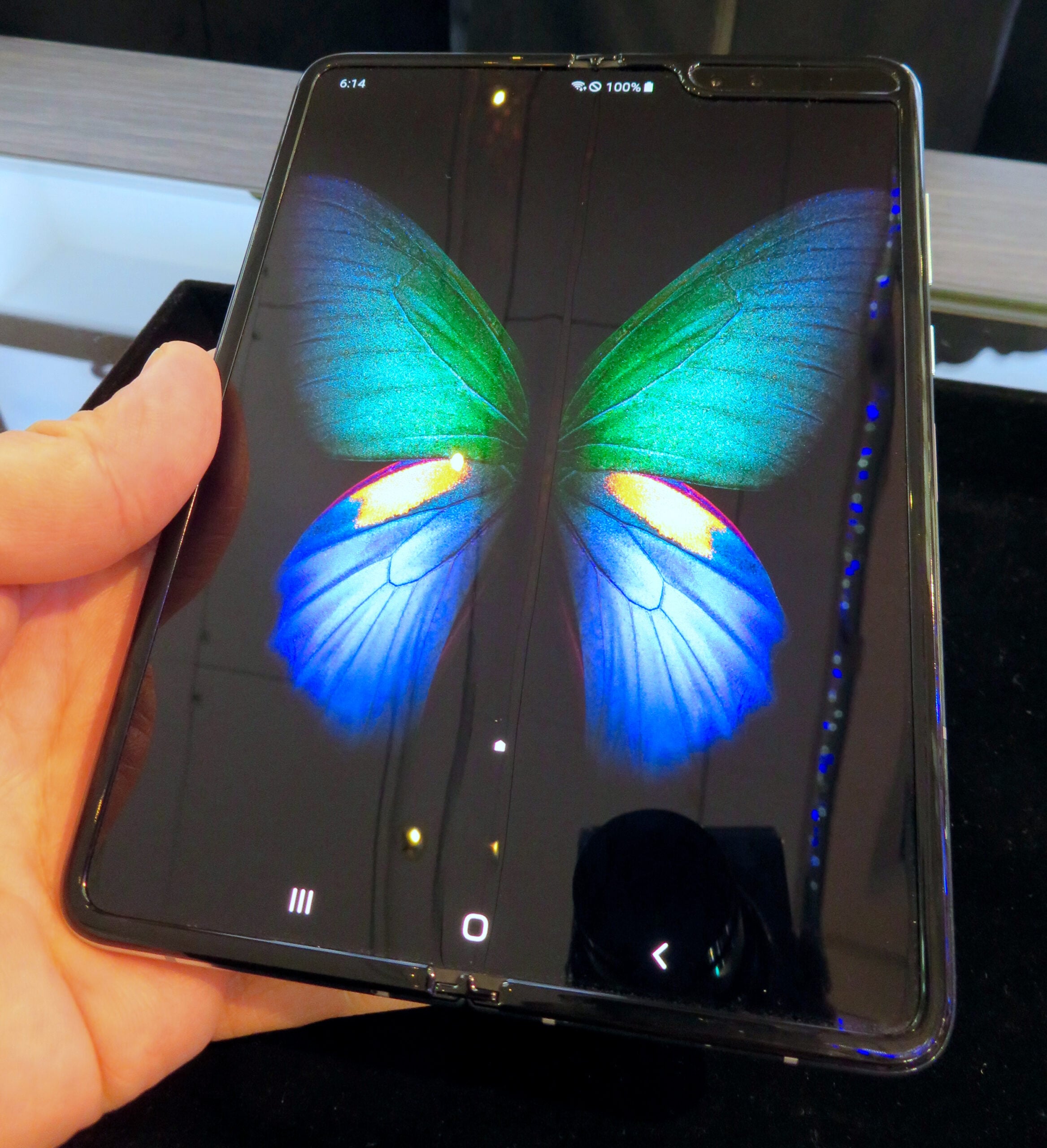 A hand holds a foldable smartphone with its screen unfolded, displaying a colorful butterfly graphic with green, blue, and yellow wings. The phone's interface is visible at the bottom of the screen.
