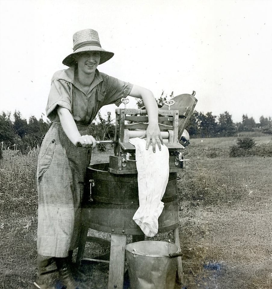A woman wearing a straw hat and checkered dress uses a hand-crank washing machine with a wringer outdoors, smiling as she works. A bucket sits below, and trees and open fields are visible in the background.