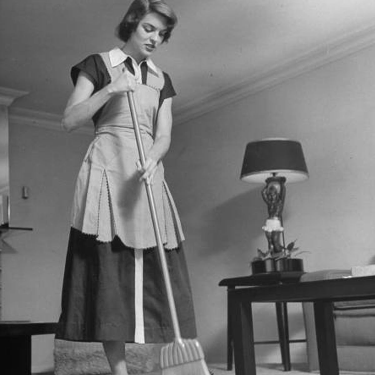 A woman in a dress and apron sweeps the floor with a broom in a tidy living room, which features a side table with a lamp and small plant.