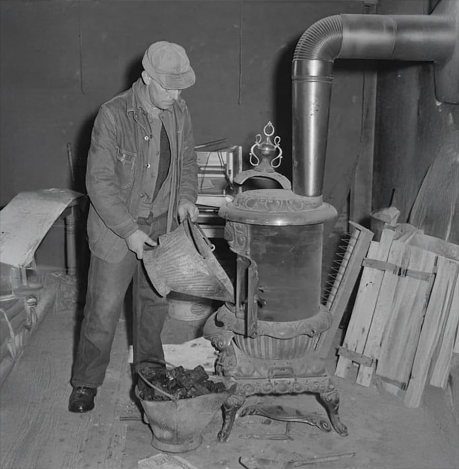 A man wearing a cap and jacket adds coal to a cast iron wood stove in a cluttered room with wooden planks and a metal pipe chimney visible.