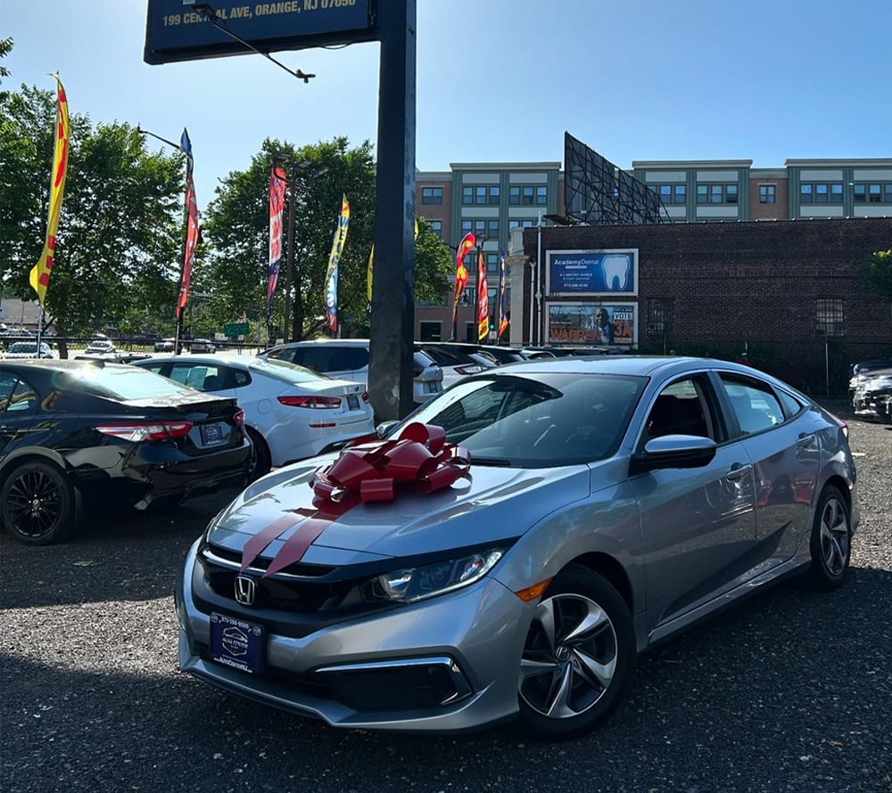 A silver Honda sedan with a large red bow on the hood is parked in a car dealership lot on a sunny day, surrounded by other vehicles and colorful flags.