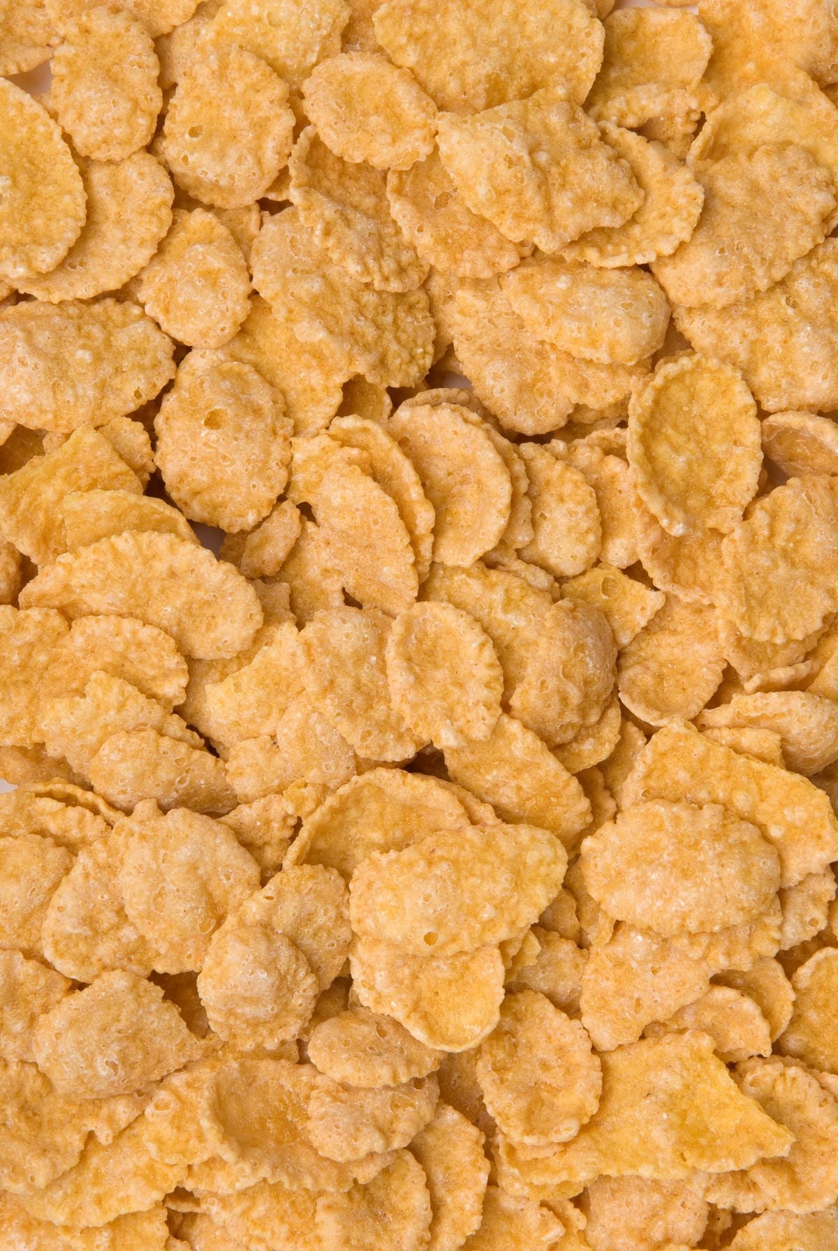 Close-up view of a pile of golden, crunchy corn flakes cereal, showing their rough texture and irregular, oval shapes.