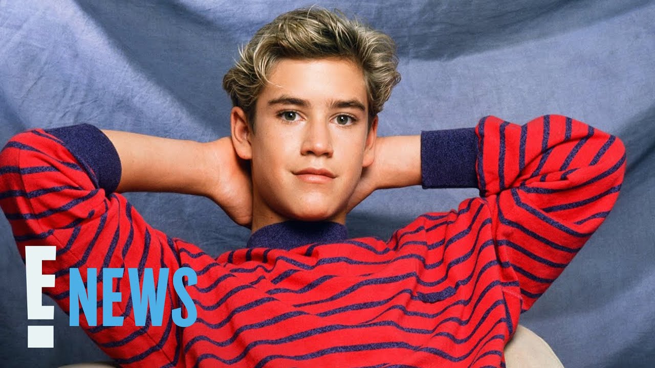A young person with short, wavy blond hair and a red striped sweater leans back with hands behind their head, posing in front of a blue backdrop. The "E! NEWS" logo appears in the bottom left corner.