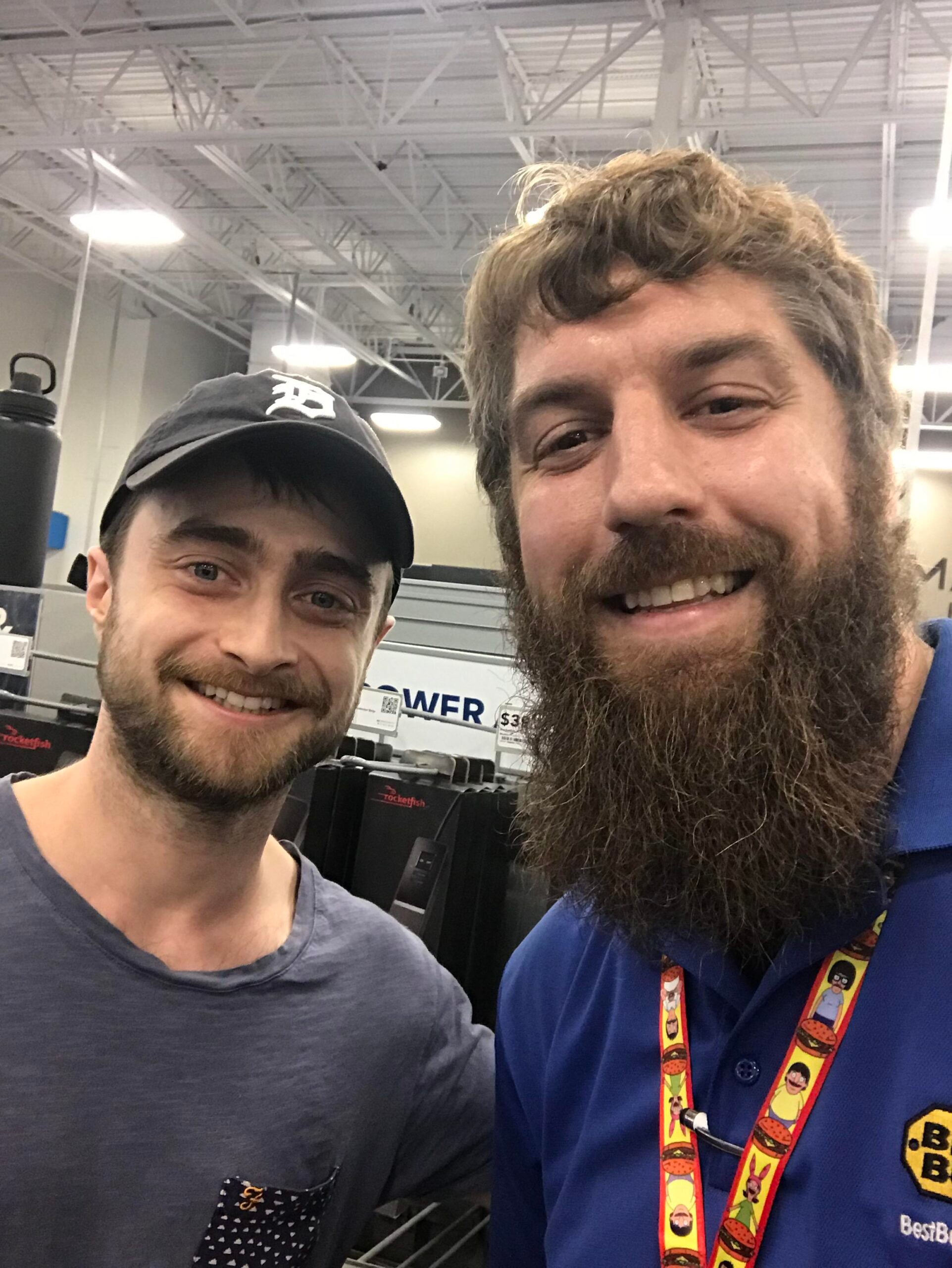 Two men smile for a selfie inside an electronics store. One wears a dark cap and t-shirt, the other has a beard, wears a blue Best Buy shirt and colorful lanyard. Store shelves and electronics are visible in the background.