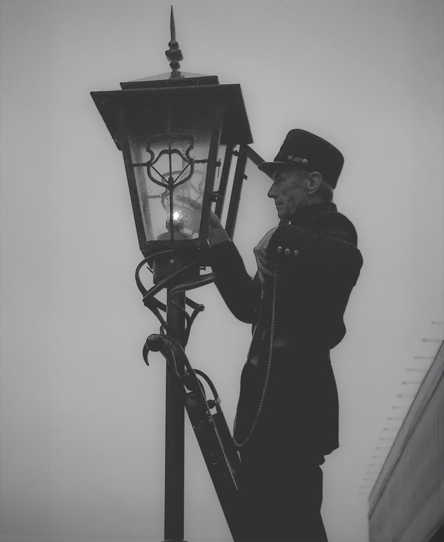 A person in uniform stands on a ladder, lighting or repairing a vintage streetlamp against a foggy or overcast sky. The image is in black and white, creating a nostalgic, historical atmosphere.