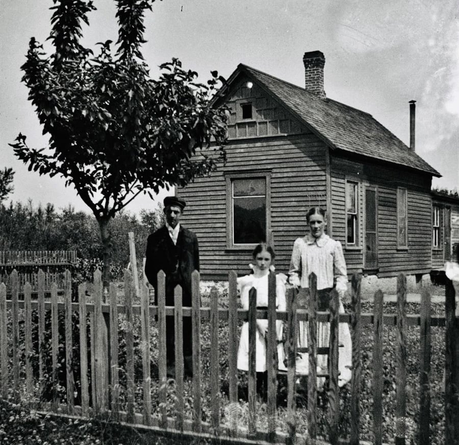 Black-and-white photo of three people—an adult man, a young girl, and an adult woman—standing behind a wooden picket fence in front of a small wooden house with a chimney and a tree nearby.
