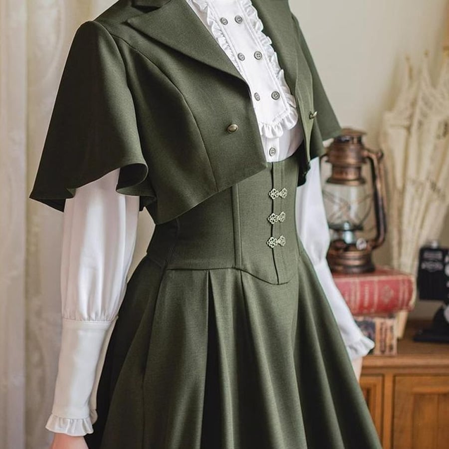 A mannequin dressed in vintage-inspired clothing: a white ruffled blouse with puffed sleeves and an olive green, high-waisted, pleated dress featuring decorative buttons and a short cape-style jacket. An antique lantern and books are in the background.