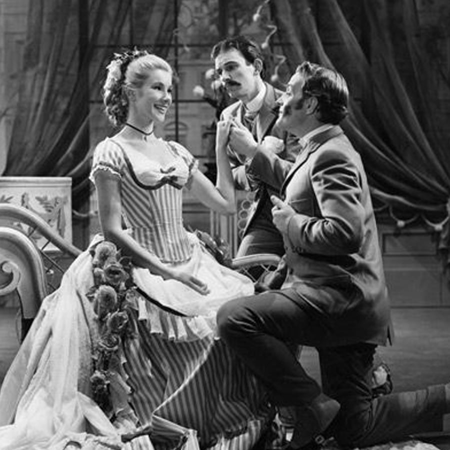 A woman in a Victorian-style dress sits on a bench, smiling, while two men in suits kneel and gesture toward her. The scene appears theatrical and set in an ornate room with draped curtains.