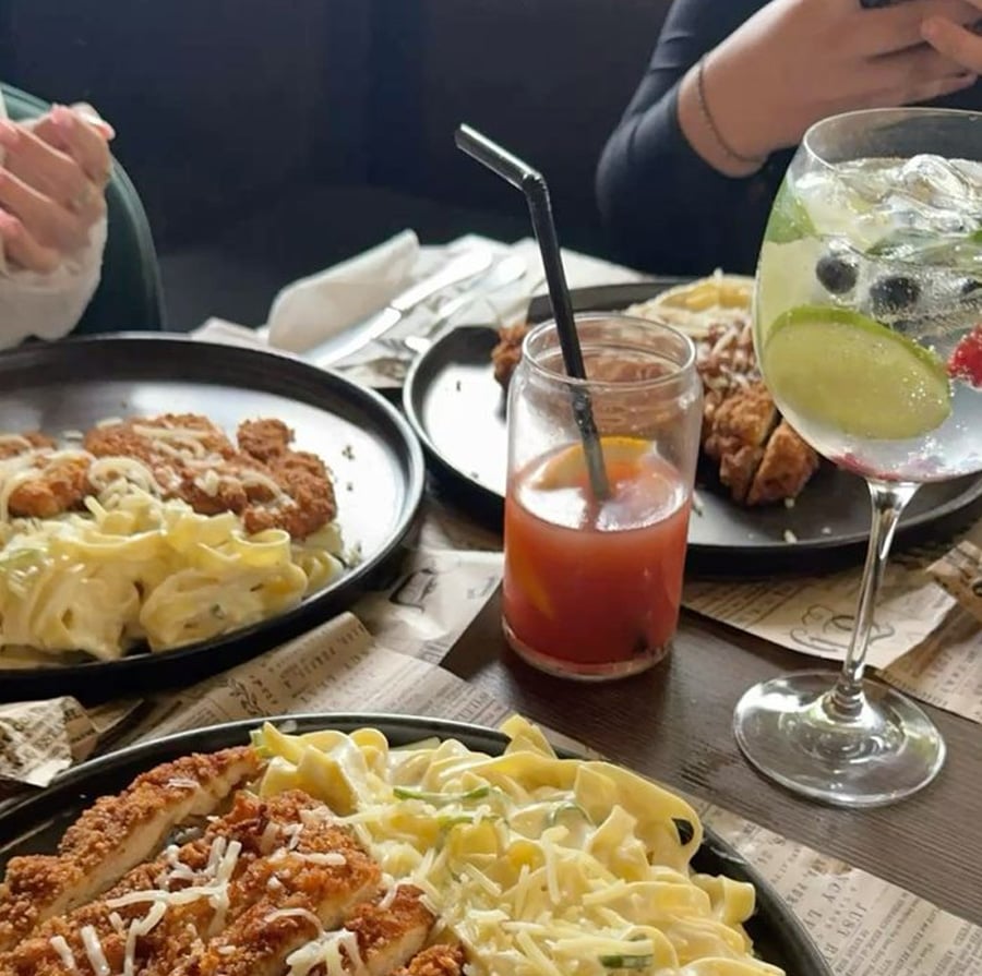 Three plates of creamy pasta topped with crispy chicken are on a table. There is also a glass of red drink with a straw and a clear cocktail with lime, berries, and ice. Two people’s hands are visible near the food.