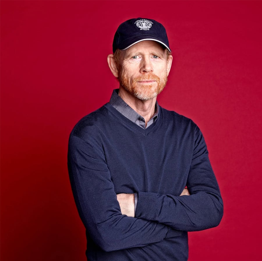 A middle-aged man with a red beard, wearing a navy blue sweater, collared shirt, and a dark baseball cap, stands with arms crossed against a solid red background.