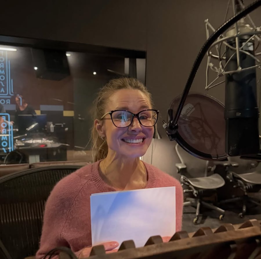 A woman wearing glasses and a pink sweater smiles while holding a piece of paper in front of a microphone in a recording studio. Audio equipment and another person are visible in the background.