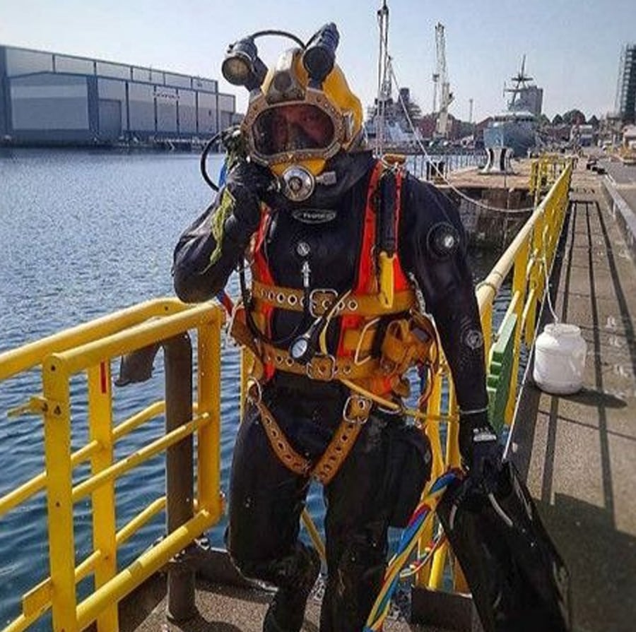 A diver in a heavy-duty suit and helmet stands on a dock near water, holding a hose and equipment. Industrial buildings, cranes, and ships are visible in the background.