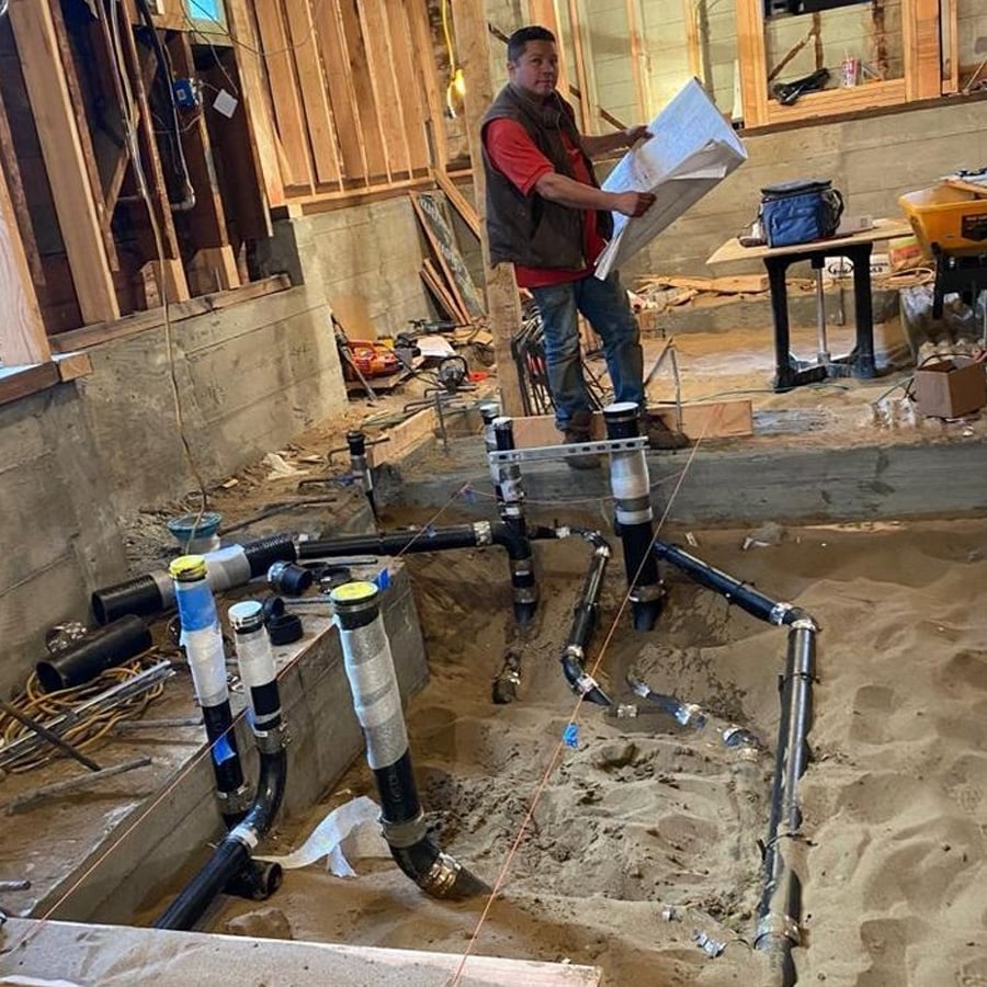 A construction worker holding blueprints stands in a partially built room with exposed wooden framing and concrete walls, inspecting newly installed black plumbing pipes set in sand.