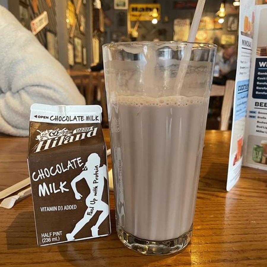 A glass of chocolate milk with a straw sits on a wooden table beside an open carton labeled "Hiland Chocolate Milk." Restaurant menus and blurred diners can be seen in the background.