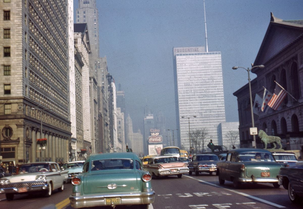 A busy city street from the 1950s or 1960s filled with vintage cars, tall buildings, American flags, and the Art Institute of Chicago with its lion statues visible on the right.