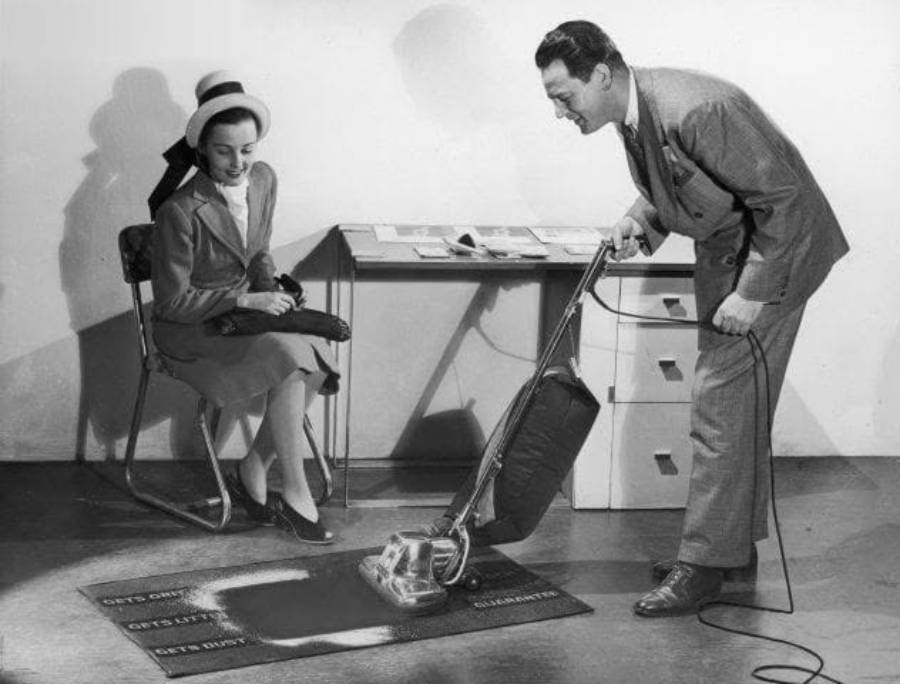 A man in a suit demonstrates a vacuum cleaner on a labeled carpet while a seated woman in a dress and hat watches. The labels on the carpet read "GETS DIRTY," "GETS LIFE," and "CLEANS IT.