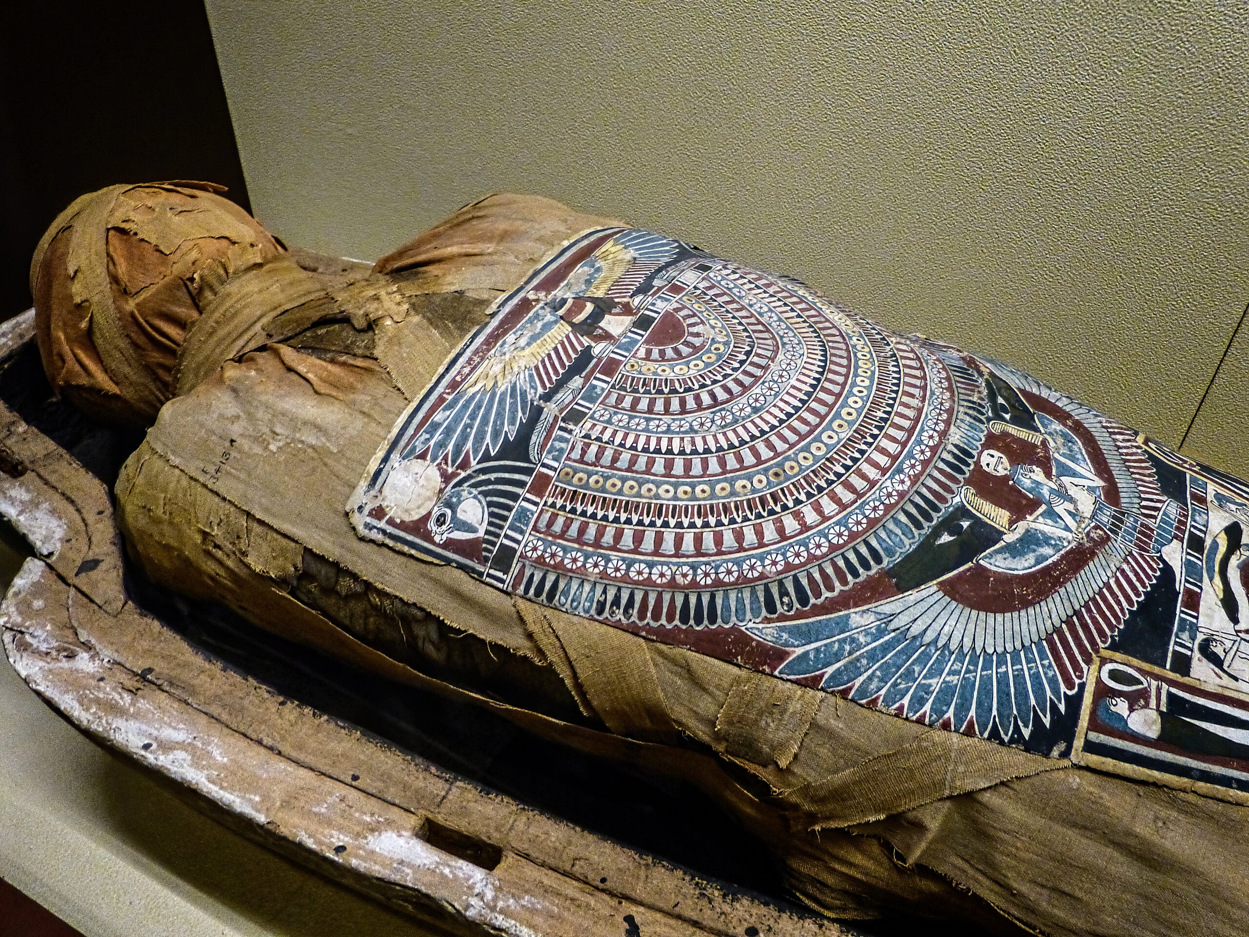 An ancient Egyptian mummy wrapped in linen, with a painted and decorated funerary mask and chest covering featuring intricate patterns and symbols, lying in a wooden coffin.
