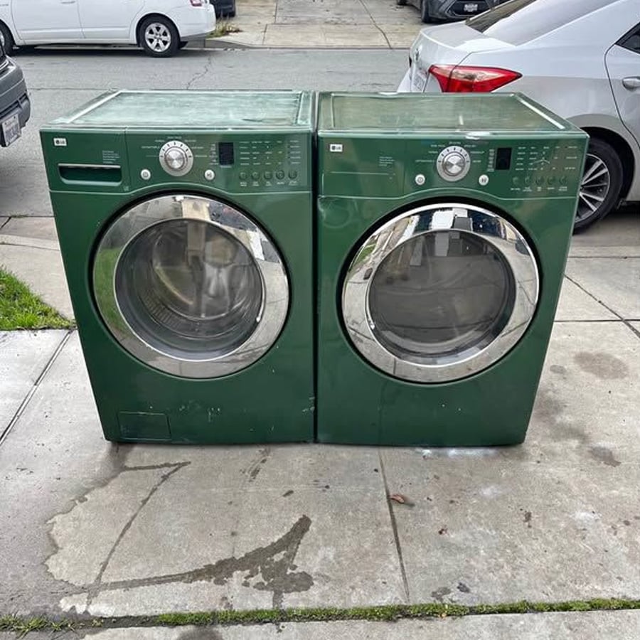 Two green front-loading washers or dryers with chrome doors are placed side by side on a sidewalk next to parked cars. The machines show signs of wear. The background includes a street and a patch of grass.