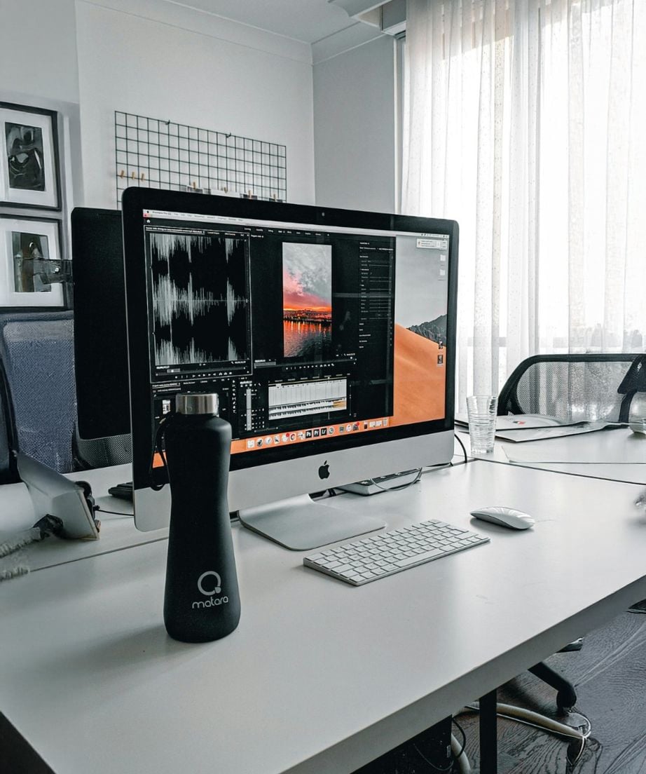 A large iMac on a white desk displays audio editing software. A black water bottle, wireless keyboard, mouse, and a glass of water are on the desk. The background shows chairs, a window with sheer curtains, and wall decor.