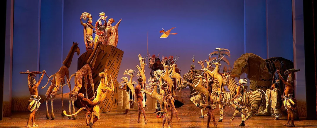 Performers in vibrant animal costumes and puppets enact a lively scene from "The Lion King" musical on stage, featuring a bright blue and orange backdrop with African-inspired scenery and props.