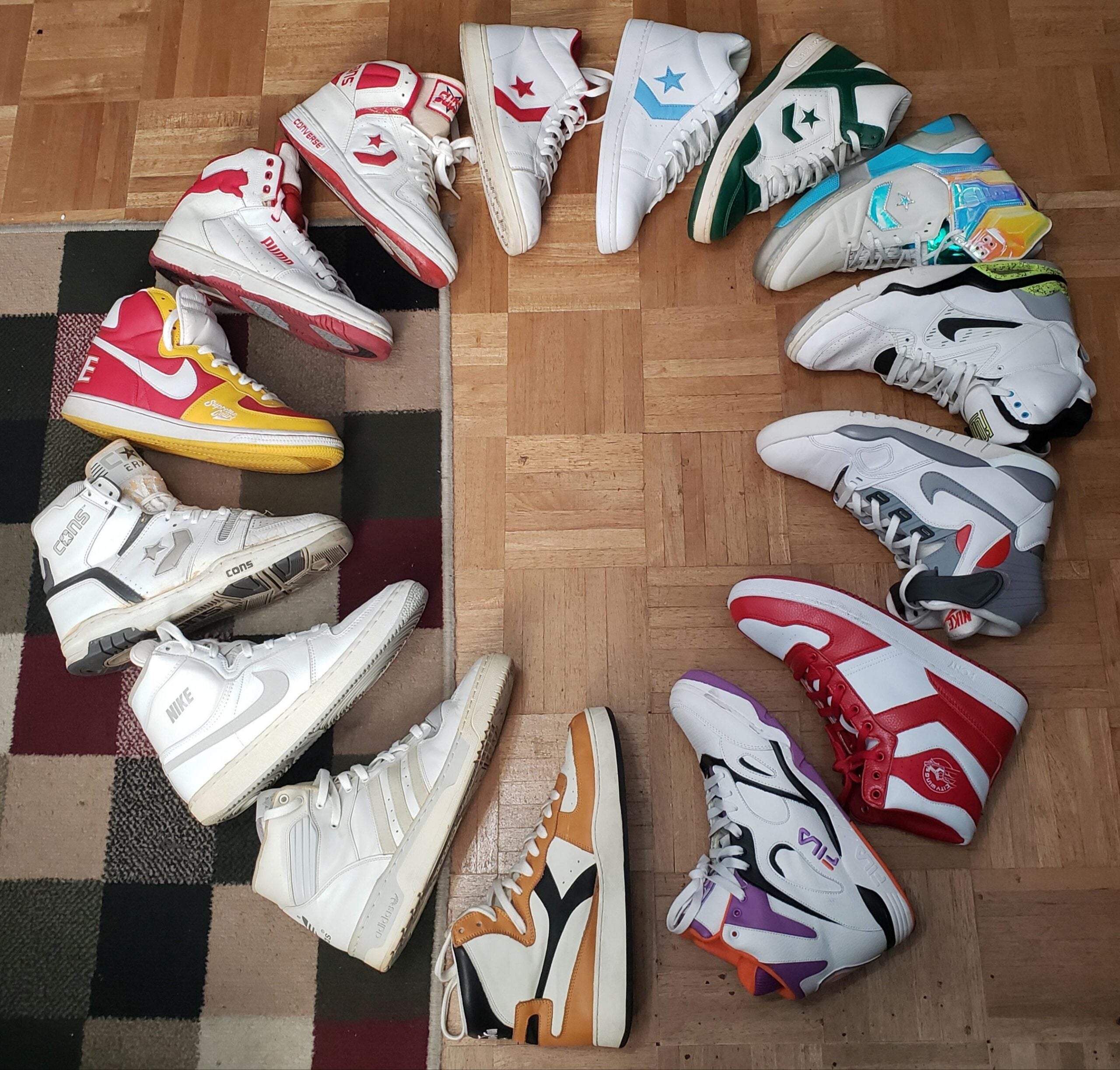 Twelve pairs of colorful high-top sneakers are arranged in a circle on a wooden floor, with each pair featuring different designs, brands, and colors. Part of a patterned rug appears in the lower left corner.