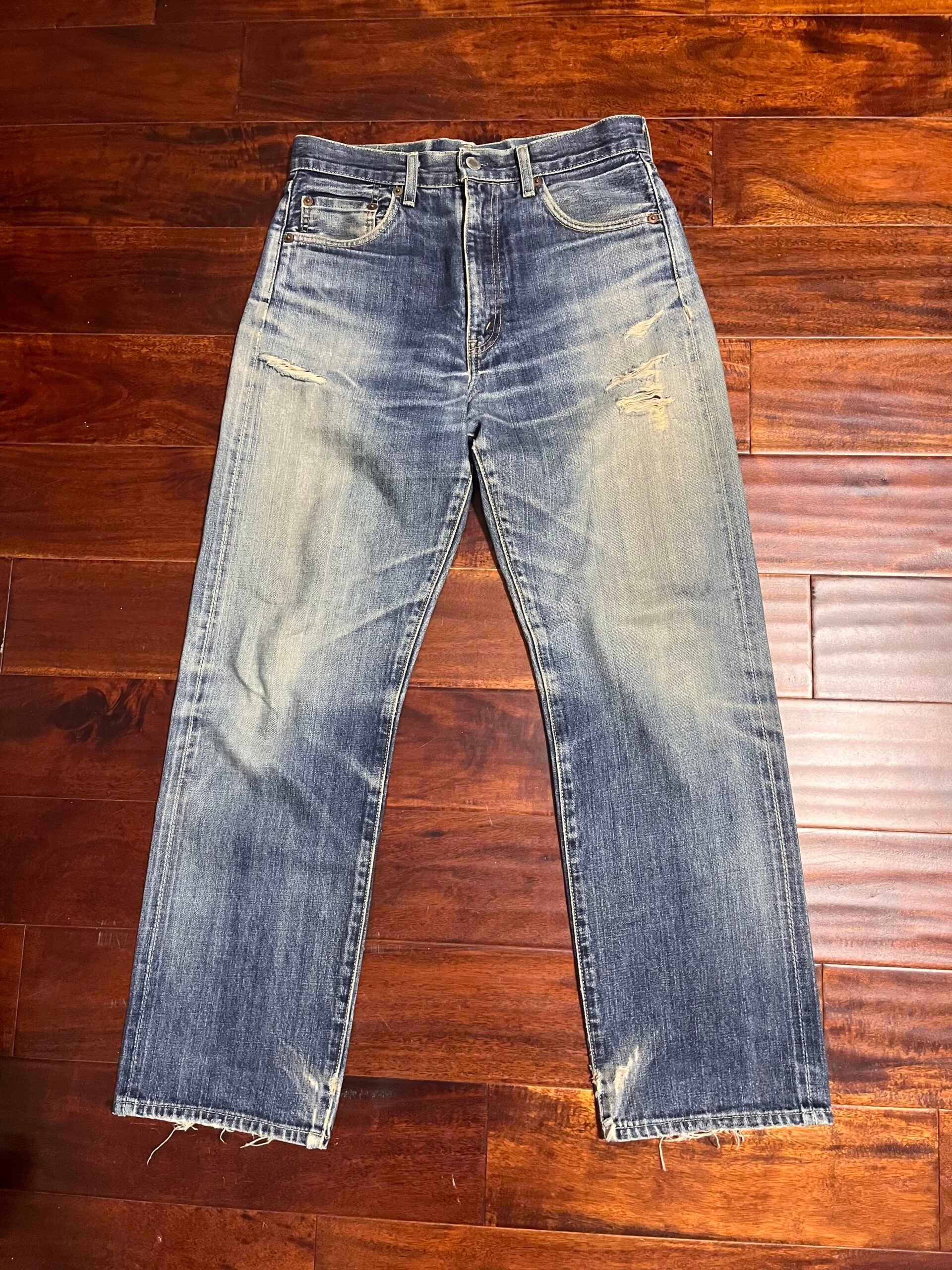 A pair of faded blue jeans with slight distressing and frayed hems is laid flat on a dark wooden floor. The jeans have a classic five-pocket design and visible wear marks.