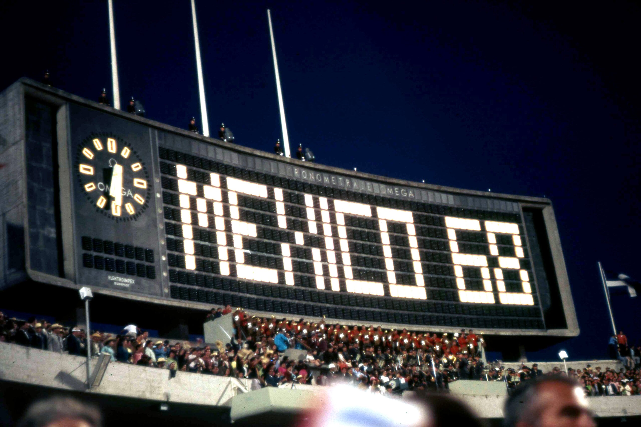 A large stadium scoreboard displays "MEXICO 68" in bright lights with a clock on the left side. A crowd is gathered below the scoreboard at night.