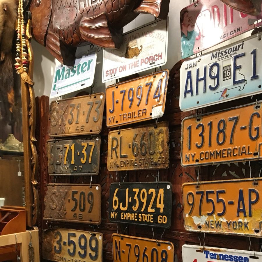 A collection of vintage license plates from various states, including New York and Missouri, displayed on a wall among eclectic items like a carved fish and a "Stay on the Porch" sign.