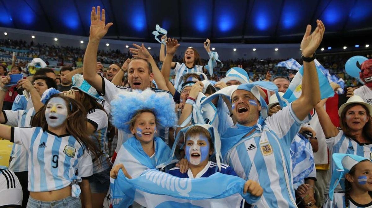 A crowd of enthusiastic Argentina soccer fans, many wearing white and blue jerseys, face paint, wigs, and waving flags, cheer excitedly in a stadium filled with spectators.