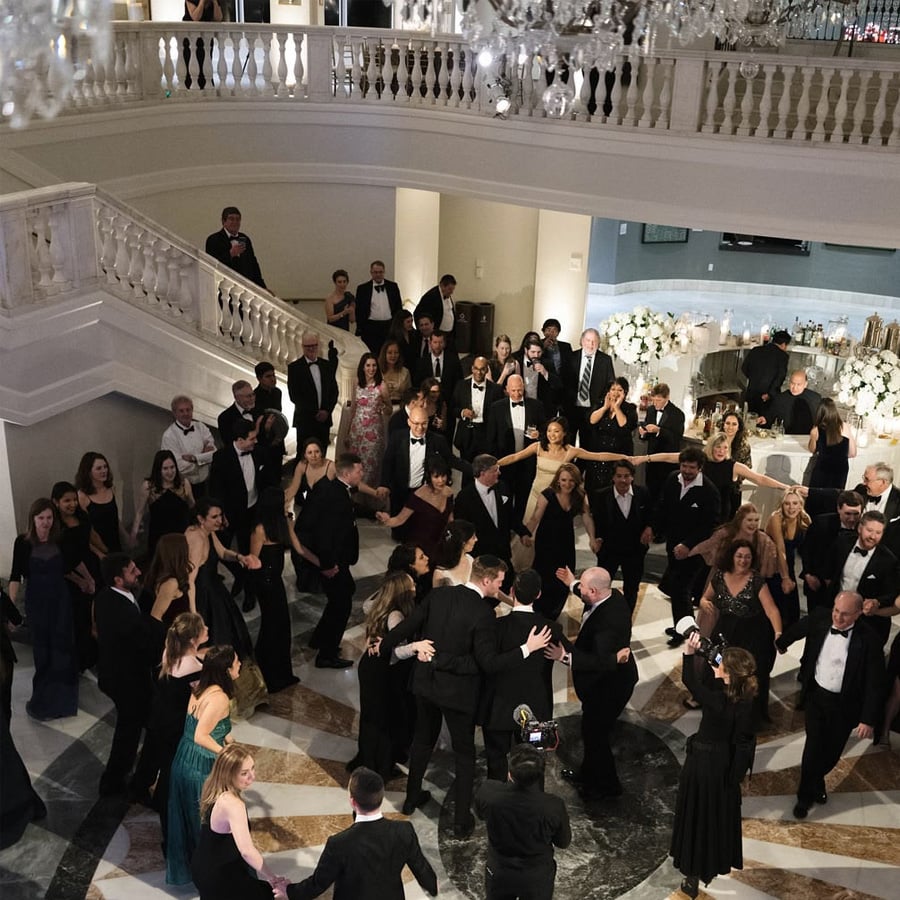 A large group of people dressed in formal evening wear dance in a circle on a marble floor in an elegant ballroom with white railings and chandeliers. Some people watch from a balcony above.