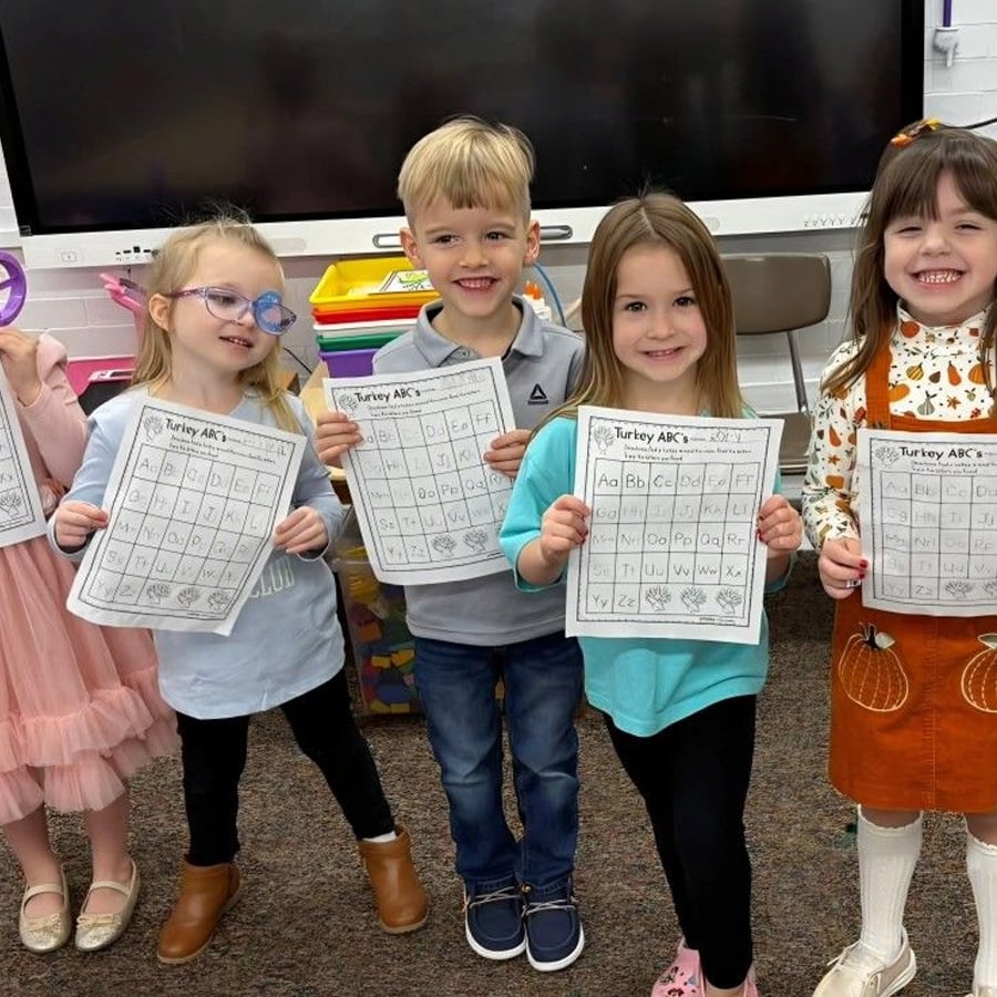 Five young children stand in a classroom, smiling and holding up completed "Turkey ABC's" worksheets showing the alphabet. A large monitor and classroom supplies are visible in the background.