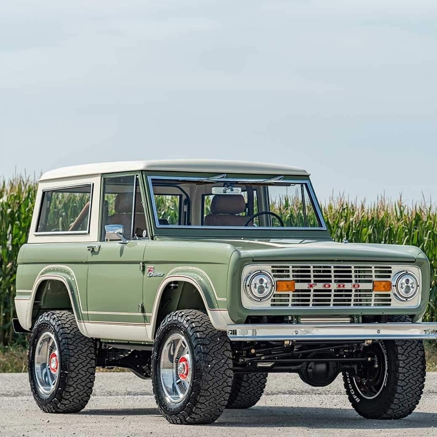A classic, light green Ford Bronco with a white roof is parked on a dirt road in front of a tall cornfield under a cloudy sky.