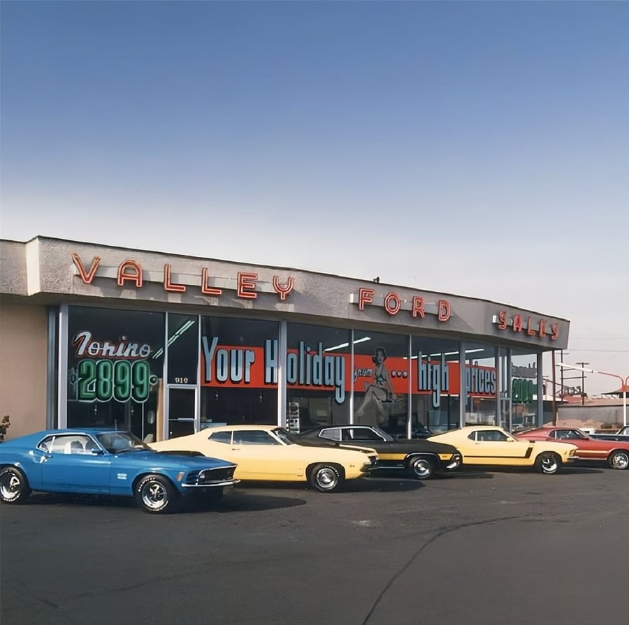 A vintage Ford dealership with neon signs displays classic cars, including blue, yellow, and orange models, parked in front of large glass windows showcasing “Your Holiday Headquarters.”