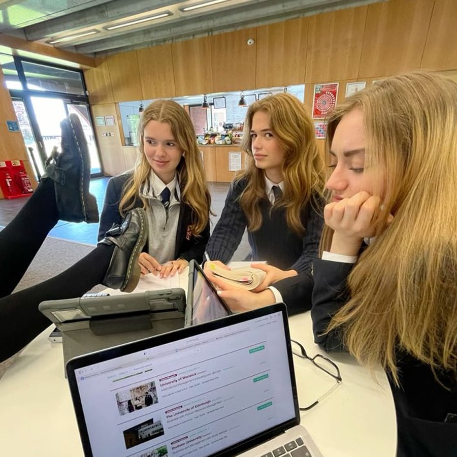 Three teenage girls in school uniforms sit at a table in a classroom, with laptops and books in front of them. One girl has her feet up on the table, while the others look on with amused and thoughtful expressions.