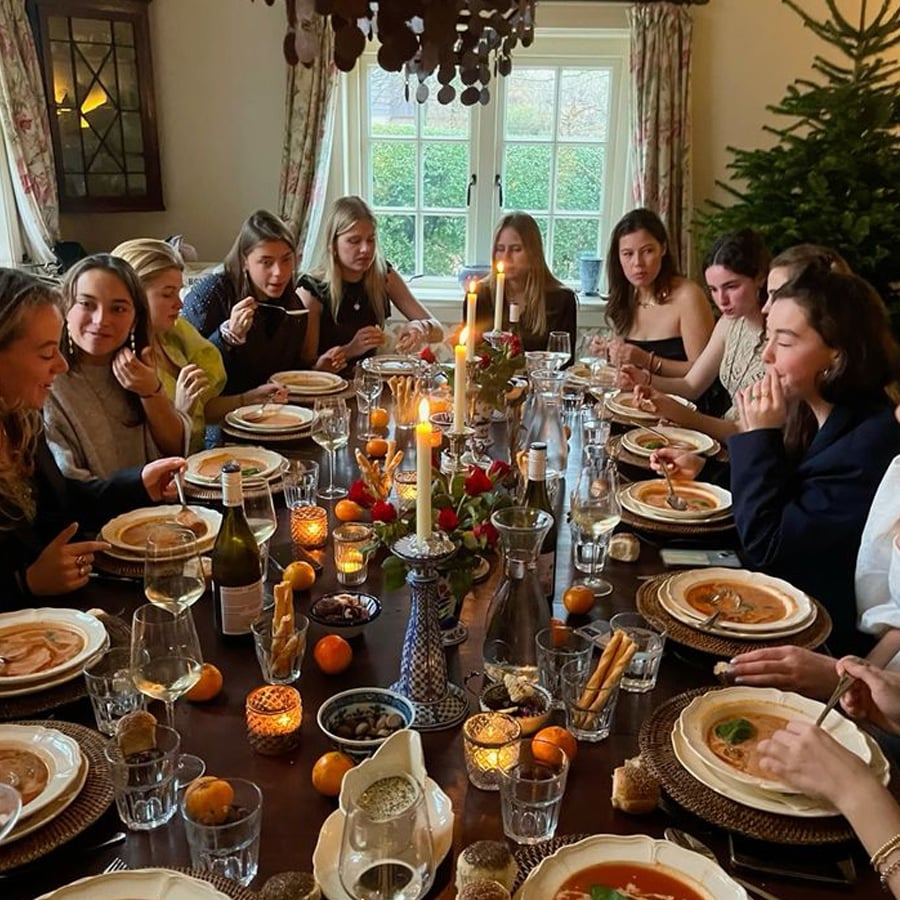 A group of twelve people sit around a candlelit dining table set with plates, glasses, and festive decorations, sharing a meal in a cozy, warmly lit room with a large window and a Christmas tree in the background.