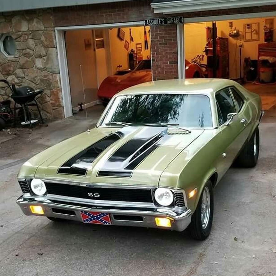 A vintage olive green Chevrolet Nova SS with black racing stripes is parked in a driveway. The car has a Confederate flag license plate. An open garage with another classic car and tools is visible in the background.