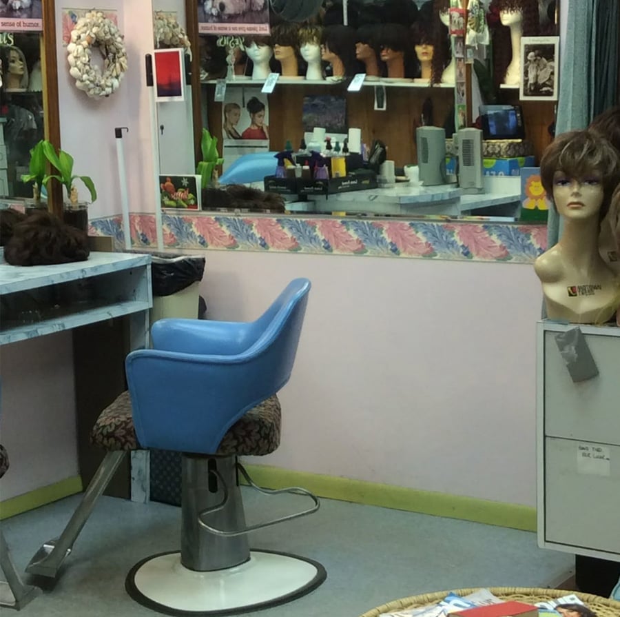 A wig shop interior with a blue salon chair, a mirror reflecting shelves of mannequin heads with wigs, hair products on a counter, and a mannequin head on a filing cabinet in the foreground.