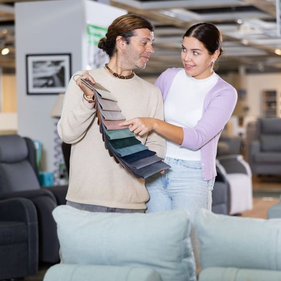 A man and woman in a furniture store look at a fabric swatch book, discussing and pointing at different colors, with sofas and chairs visible in the background.