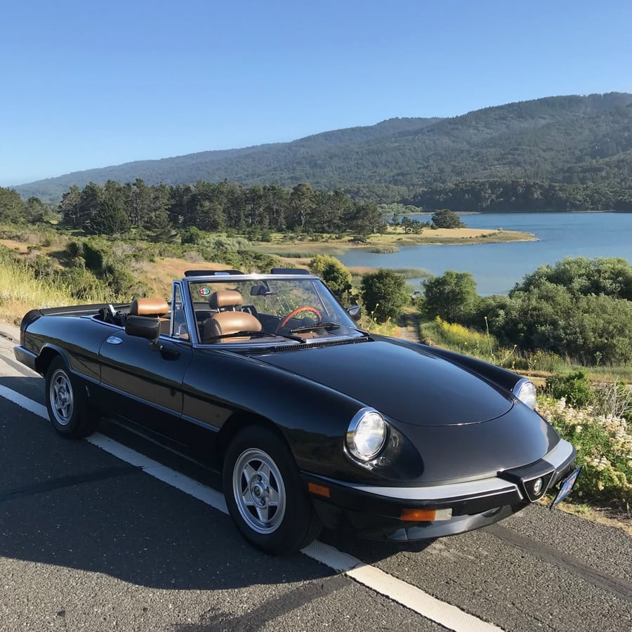 A black vintage convertible car is parked on a road next to a scenic lake, with green hills and trees in the background under a clear blue sky.