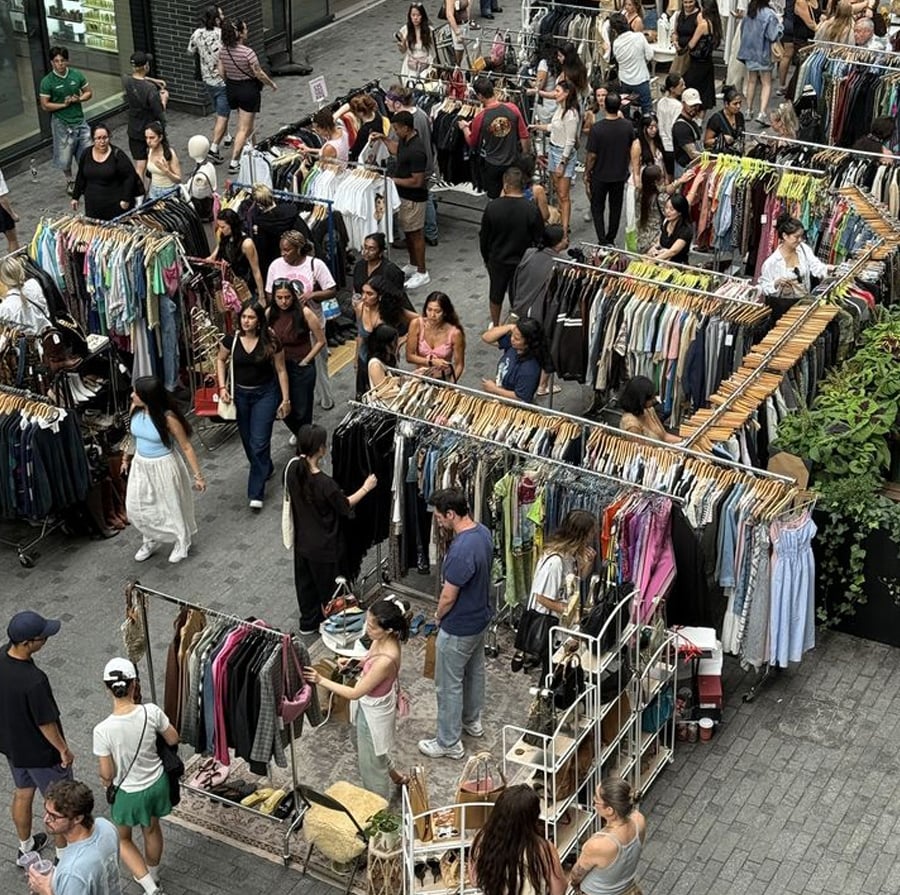 A busy outdoor market with people browsing racks of clothes and accessories, surrounded by greenery and buildings. The market has a lively atmosphere with shoppers exploring different stalls.