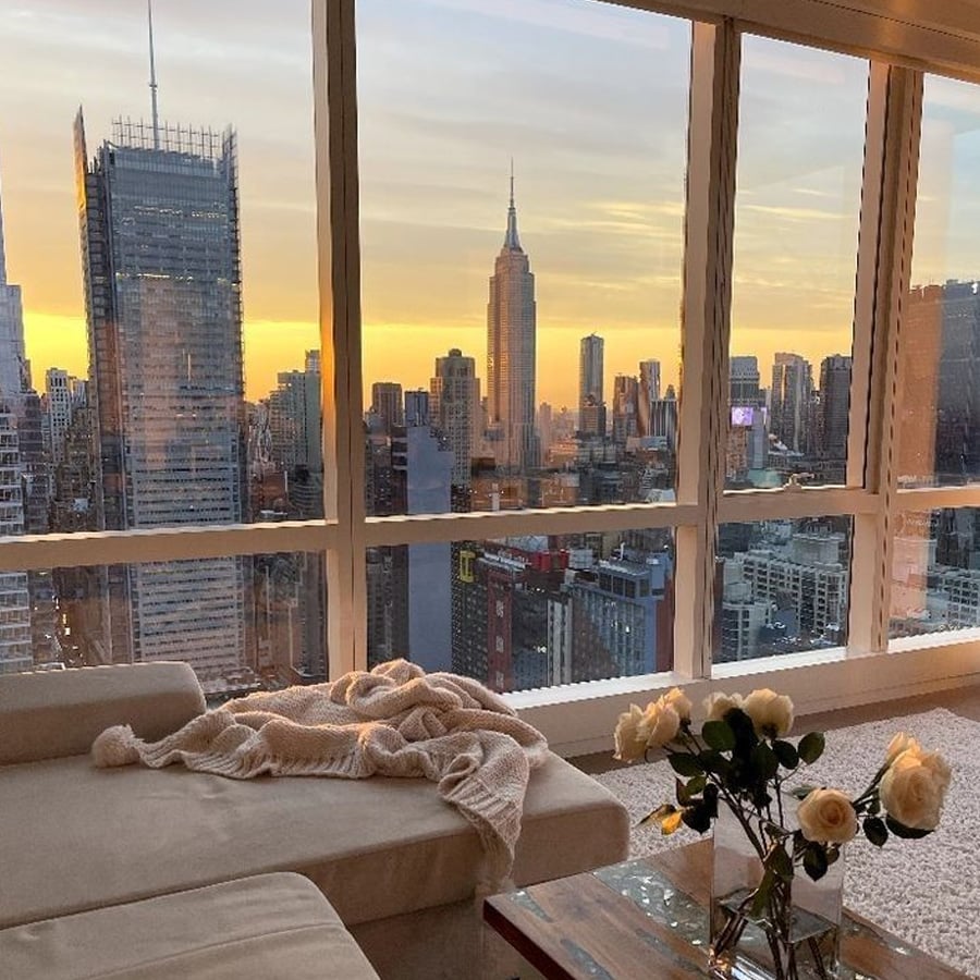 A cozy living room with a beige sofa and white roses on a table overlooks a city skyline at sunset, featuring the Empire State Building and other skyscrapers through large floor-to-ceiling windows.