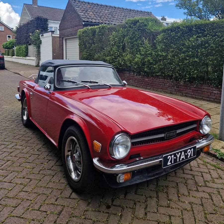 A classic red convertible car with a black soft top is parked on a brick street in a residential neighborhood, with houses and greenery in the background.
